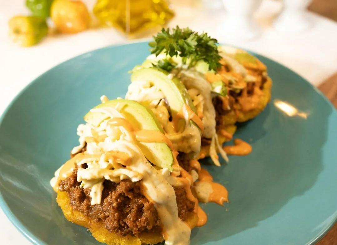 Two fried arepas topped with pulled meat, cole slaw, avocado slices and sauce, on a teal plate.