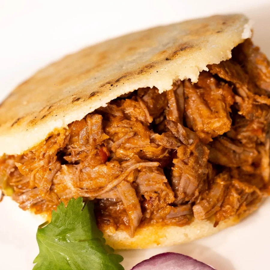 A griddled arepa split open and filled with shredded beef, garnished with cilantro and red onion.