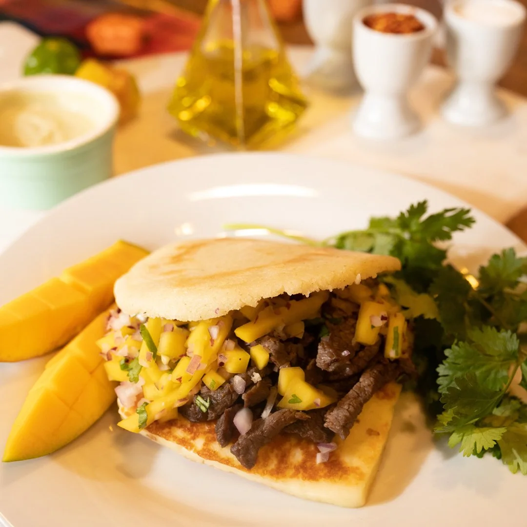 An arepa filled with sliced steak and mango pico de gallo, with fried sweet plantains and cilantro alongside.