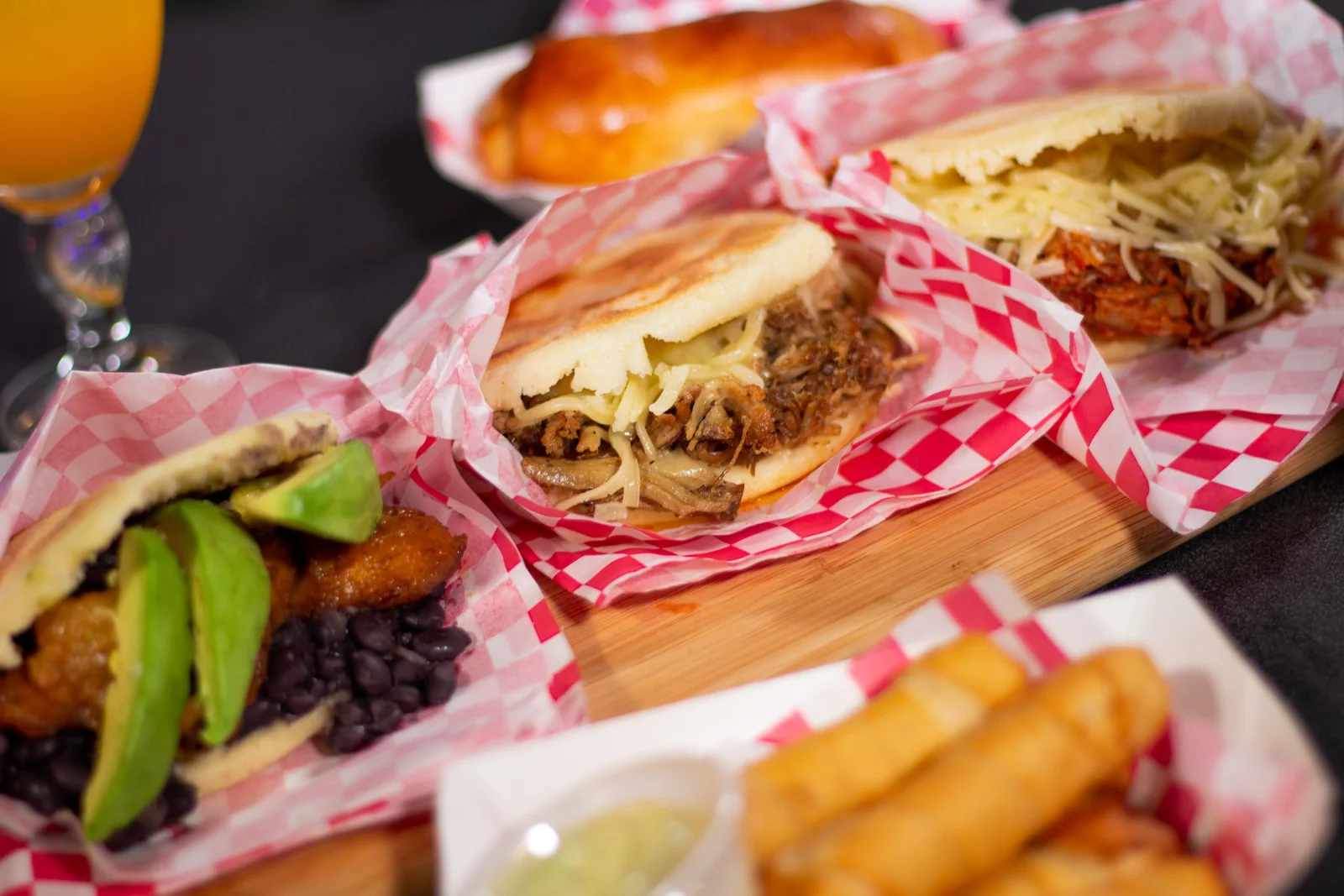 An arepa filled with pulled pork and shredded cheese in a checkered paper basket, with tequeños beside it.
