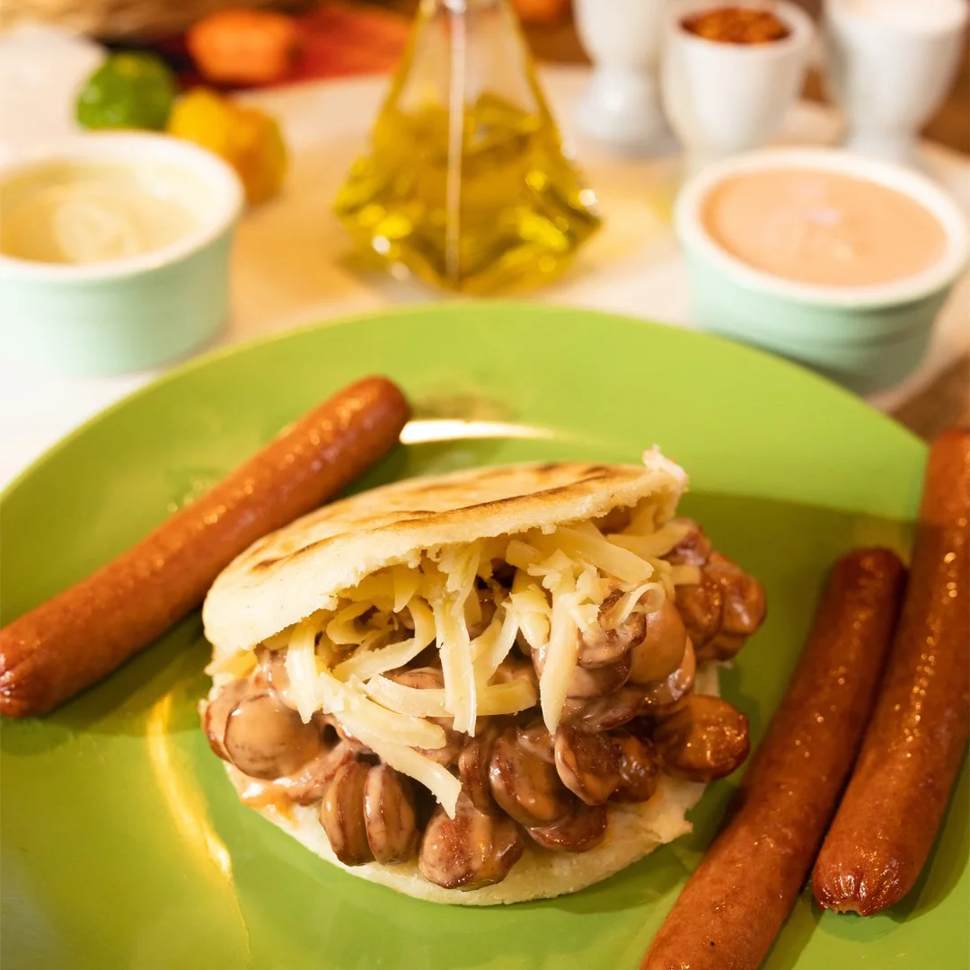 An arepa filled with beans and shredded white cheese on a green plate, with two sausages beside it.