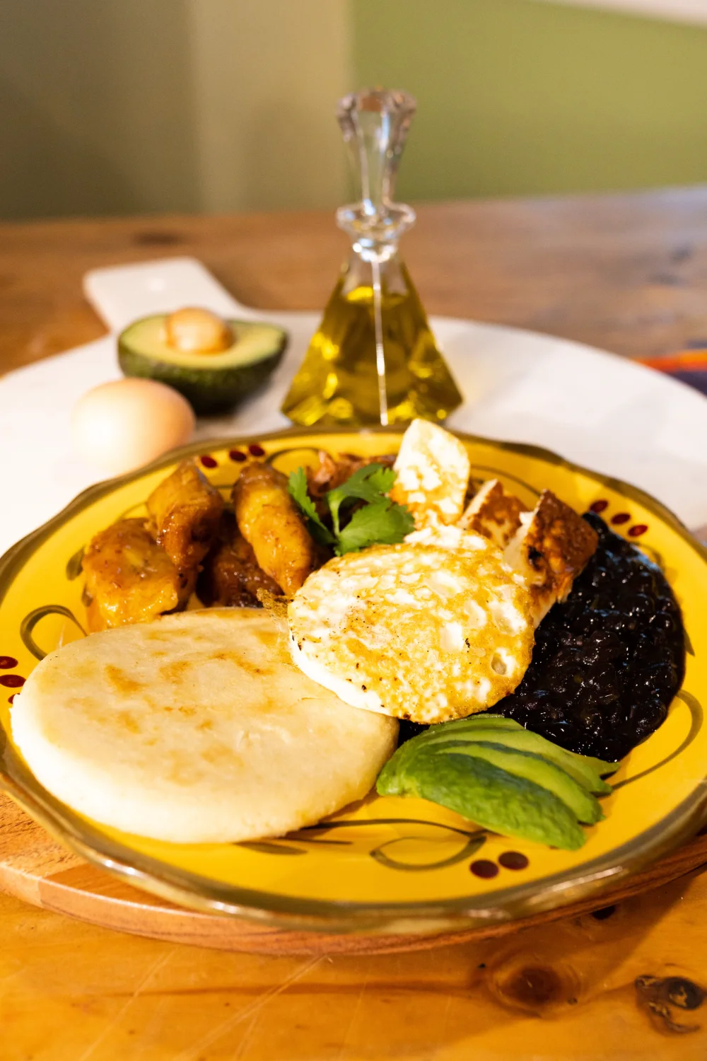 An arepa with black beans, fried plantain, avocado, a fried egg and shredded beef on a yellow patterned plate.