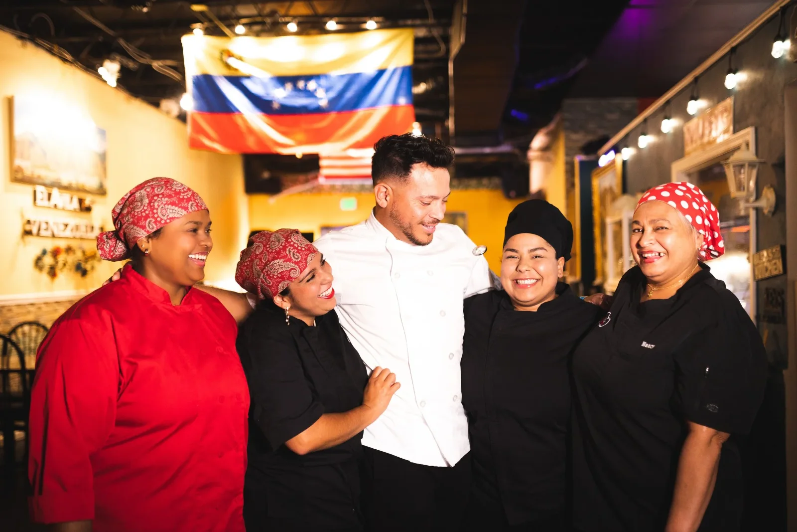 Five members of the kitchen team standing arm in arm in the dining room, four in red headscarves and one in a white chef's coat.