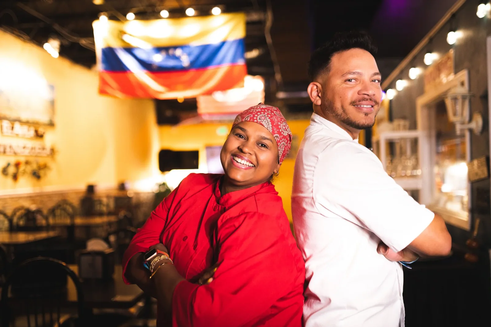 Two cooks standing back to back and smiling, one in a red chef's coat, one in a white one.