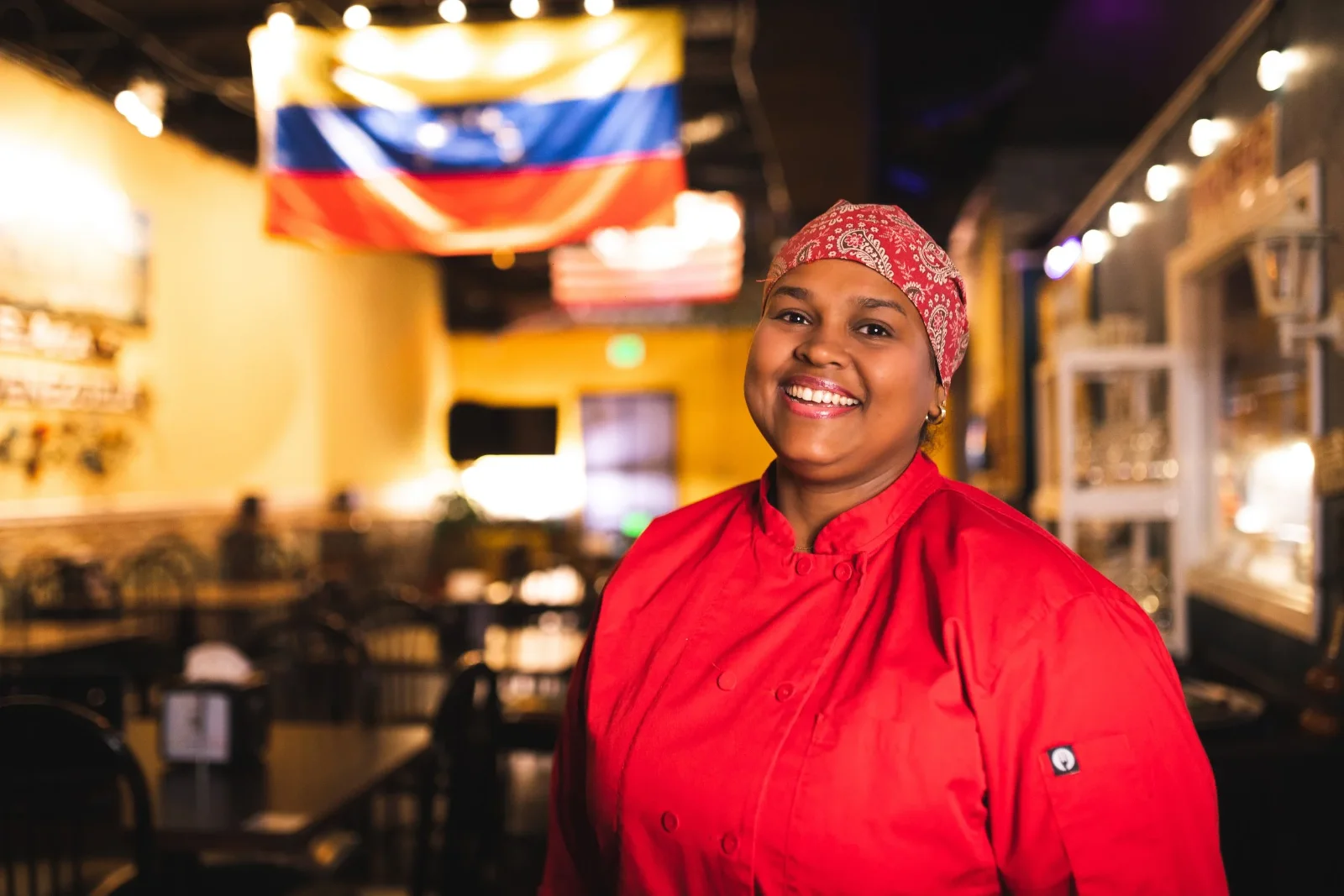 A cook in a red chef's coat and a red headscarf smiling in the dining room, with a Venezuelan flag hanging overhead.