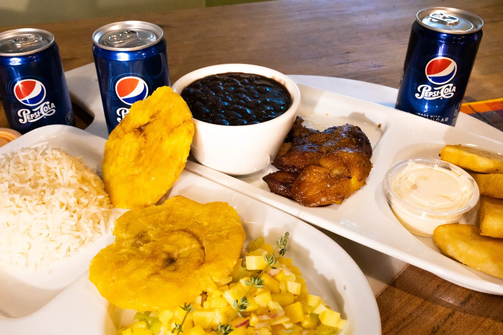A plate of fried sweet plantains, black beans, mango pico de gallo and grilled chicken, with cans of soda behind it.