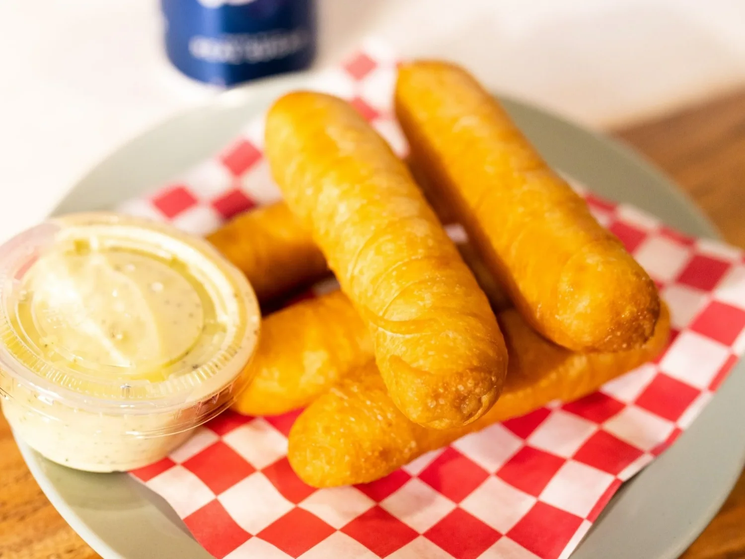 Tequeños, white cheese fingers in crisp fried dough, served on checkered paper with a cup of dipping sauce.