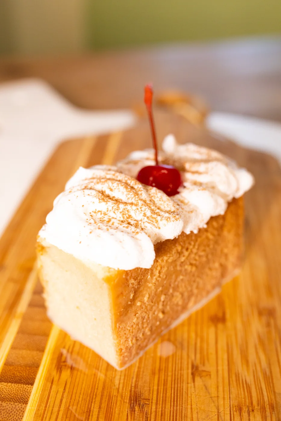 A slice of tres leches cake topped with whipped cream, cinnamon and a cherry, on a wooden board.